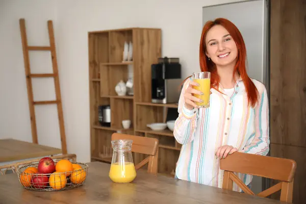Young redhead woman with glass of juice in kitchen