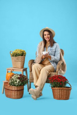 Elinde kahve ve kasımpatı çiçekleri olan genç bir kadın mavi arka planda koltukta oturuyor.