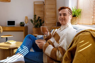 Evdeki kanepede oturan bir fincan kahveyle mutlu kızıl saçlı genç adam.