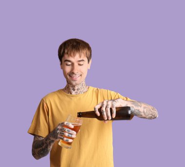 Dövmeli genç mutlu adam mor arka planda bardağa bira dolduruyor.