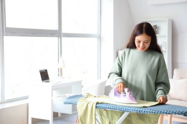 Genç Asyalı kadın evde kıyafetlerini ütülüyor.