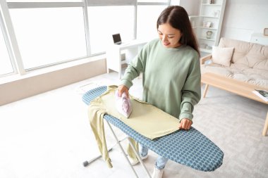 Genç Asyalı kadın evde kıyafetlerini ütülüyor.