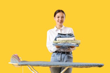 Sarı arka planda ütü masasının yanında duran temiz elbiseli mutlu Asyalı kadın.