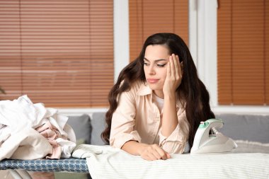 Evde çamaşır yığınıyla ütü masasına yaslanan yorgun genç bir kadın.