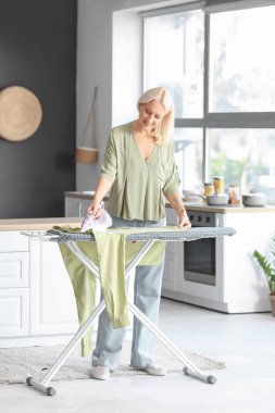 Evde kıyafetlerini ütüleyen mutlu olgun bir kadın.