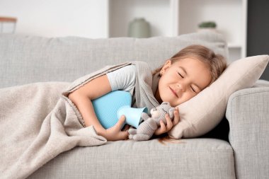 Hasta küçük kız sıcak su şişesi ve oyuncağıyla evdeki kanepede yatıyor.