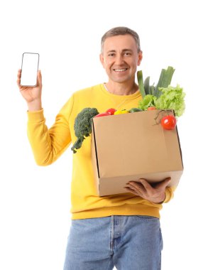 Beyaz arka planda bir kutu taze sebze ve cep telefonu olan olgun bir adam.