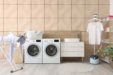 Çamaşır odasının içinde çamaşır makinesi, buhar makinesi ve ütü masası var.