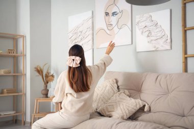 Kanepedeki genç kadın oturma odasındaki şık resimlere bakıyor.