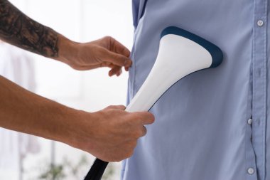 Genç adam evde mavi gömleği buharlaştırıyor.