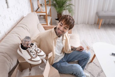 Hediye kartı ve yeni ayakkabıları olan genç dövmeli adam evdeki kanepede oturuyor.