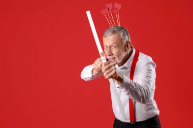 Aşk tanrısı gibi giyinmiş olgun bir adam kırmızı arka planda ok atıyor. Sevgililer Günü kutlaması