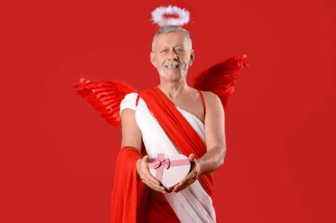 Kırmızı arka planda hediye kutusu olan Eros gibi giyinmiş olgun bir adam. Sevgililer Günü kutlaması