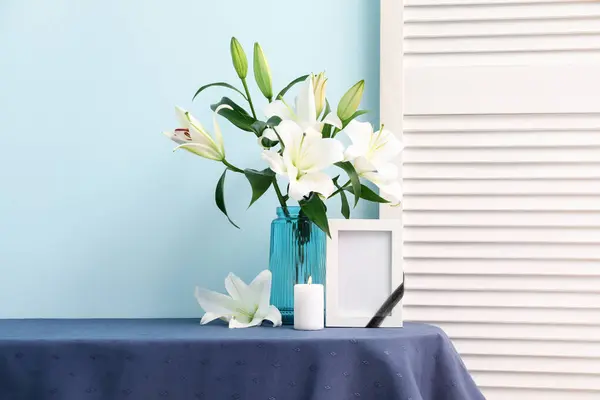 Blank funeral frame, burning candle and vase with lily flowers on table near color wall