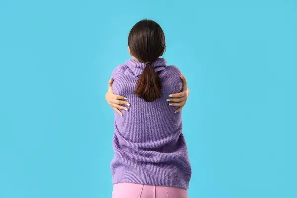 Beautiful young woman hugging herself on blue background, back view
