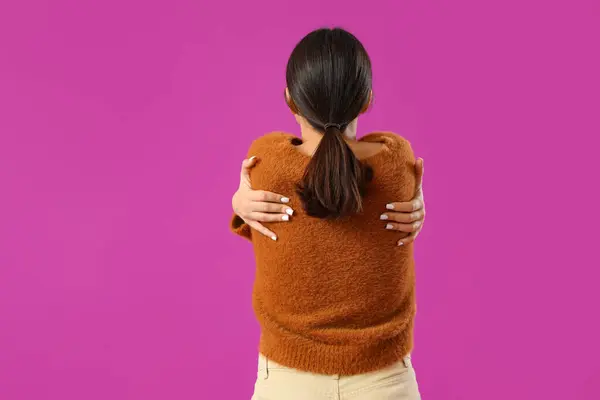 Beautiful young woman hugging herself on purple background, back view
