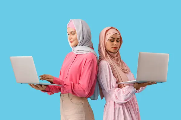 Beautiful young Muslim women in hijab with laptops on blue background