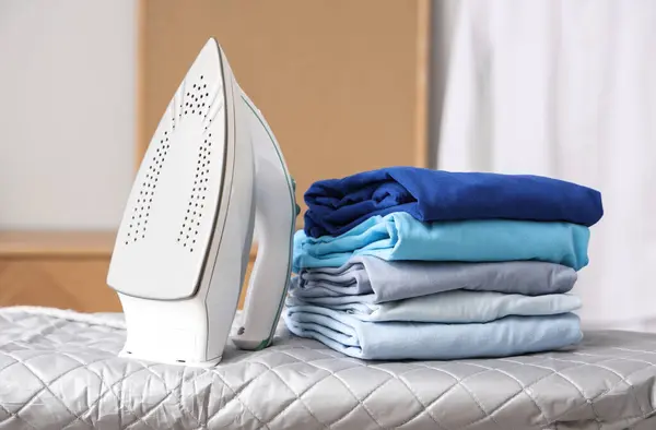 Ironing board with stack of clean clothes and modern iron at home, closeup
