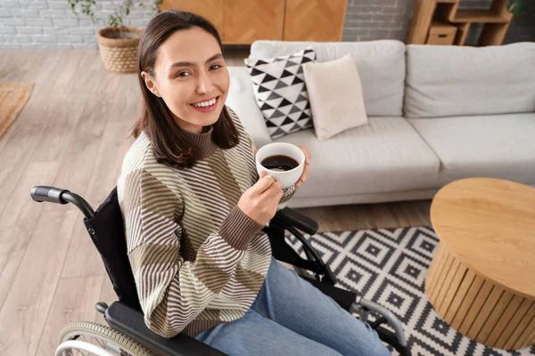 Happy young woman in wheelchair with cup of coffee at home