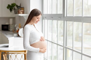 Mutfakta pencerenin yanında gülümseyen hamile bir kadın.