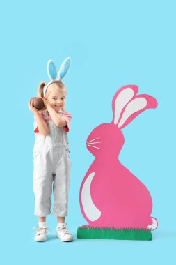 Çikolata yumurtası ve mavi arka planda kağıt tavşanla tavşan kulaklı şirin bir kız.