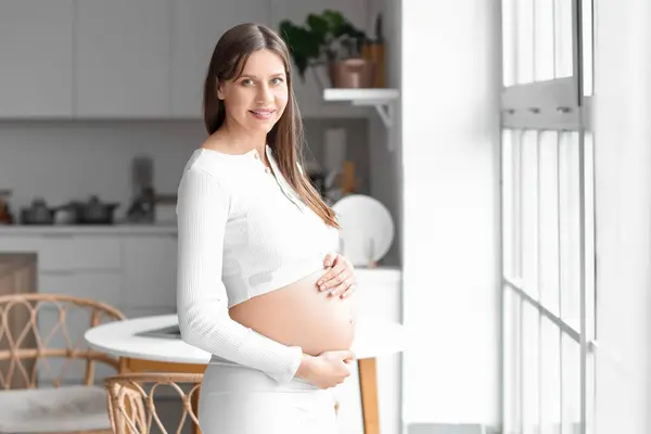 Young pregnant woman smiling near window in kitchen