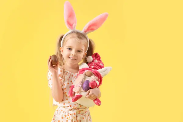 可爱的小女孩 长着小兔子耳朵 戴着巧克力复活节彩蛋和黄色背景的玩具 — 图库照片