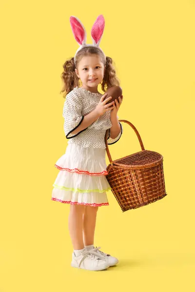 可爱的小女孩 长着小兔子耳朵 戴着巧克力复活节彩蛋和黄色背景的篮子 — 图库照片