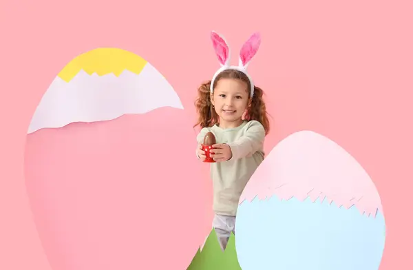 在兔子耳朵里可爱的小女孩 戴着巧克力复活节彩蛋 粉红背景的纸装饰 — 图库照片