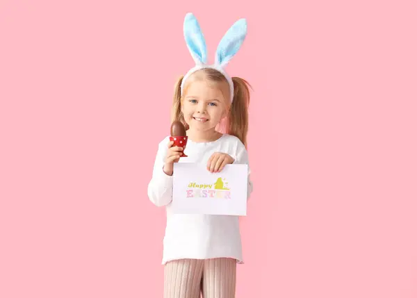在兔子耳朵里可爱的小女孩 戴着巧克力复活节彩蛋和粉色背景卡片 — 图库照片