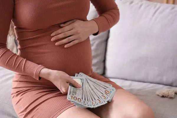 Young pregnant woman with money sitting on sofa at home, closeup. Maternity Benefit concept