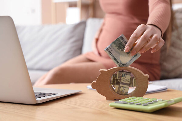 Young pregnant woman putting money into piggy bank at home, closeup. Maternity Benefit concept