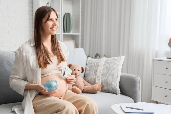 Young pregnant woman with money and piggy bank sitting at home. Maternal Benefit concept