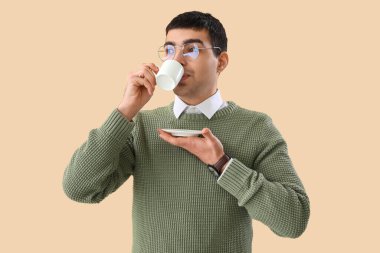 Gözlüklü genç adam bej arka planda kahve içiyor.