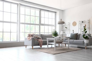 Büyük pencerenin yanında gri kanepeleri ve kahve masası olan aydınlık oturma odasının içinde.