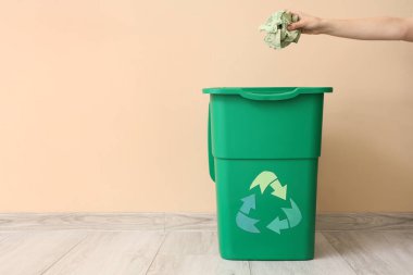 Bej duvarın yanında geri dönüşüm logosu olan çöp kutusuna buruşmuş kağıt atan bir kadın el.