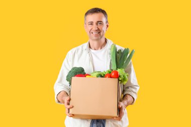 Sarı arka planda bir kutu taze sebzeli olgun adam.