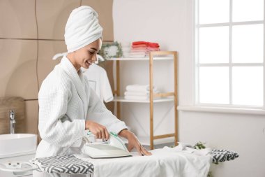 Çamaşırhanede bornozlu güzel bir kadın çamaşırları ütülüyor.