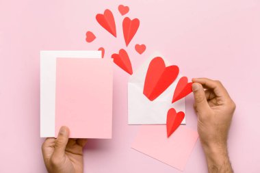 Zarflı erkek eller, boş kartlar ve pembe arka planda kırmızı kağıt kalpler. Sevgililer Günü kutlaması