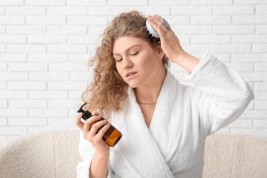 Saç derisi masajcısı ve beyaz tuğlalı arka planda bir şişe şampuanı olan güzel bir genç kadın.