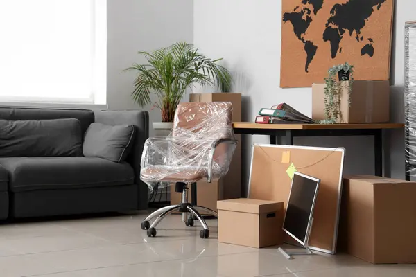 Interior of office with chair wrapped in stretch film, sofa and cardboard boxes on moving day