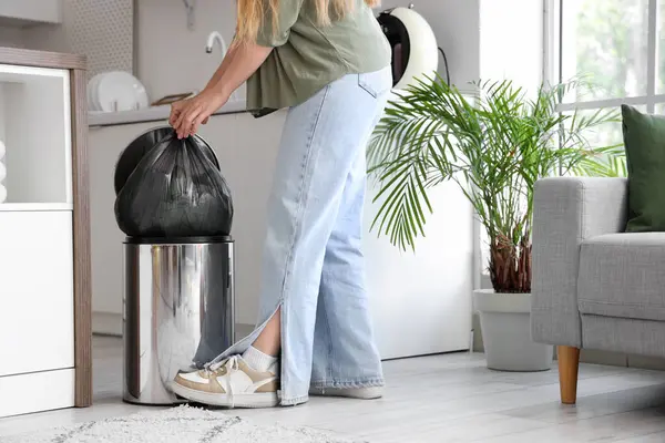 Woman throwing full garbage bug into trash bin with foot pedal in kitchen