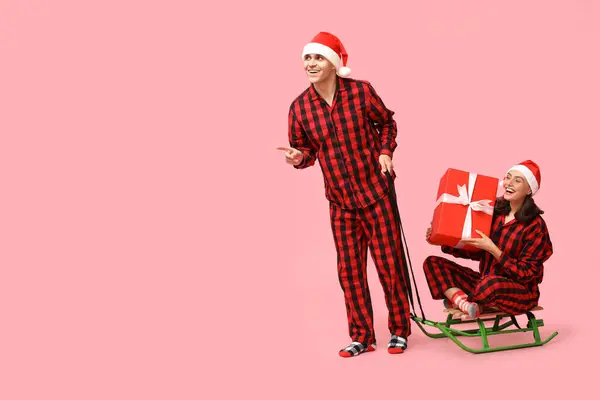 Glada Unga Par Julpyjamas Med Släde Och Gåva Rosa Bakgrund — Stockfoto