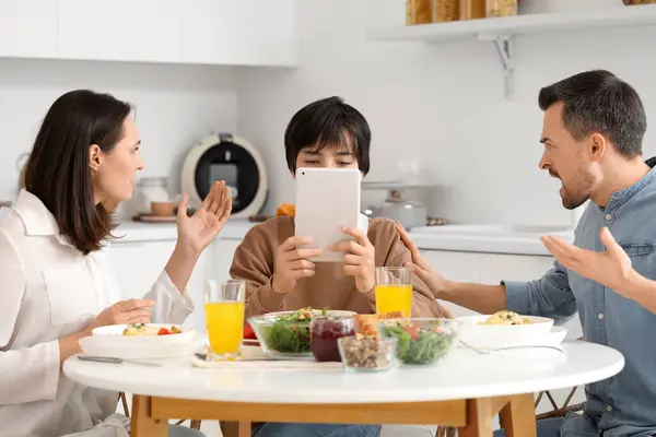 Teenage boy using tablet computer and his angry parents at table during dinner in kitchen. Family problem concept