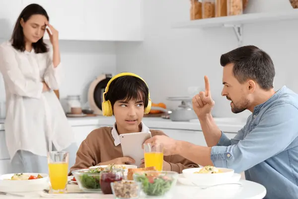 Angry father taking tablet computer away from his teenage son at table during dinner in kitchen. Family problem concept