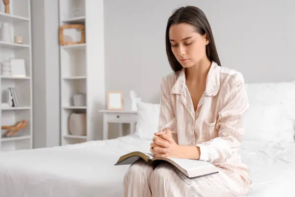 Beautiful young woman reading Bible and praying in bedroom