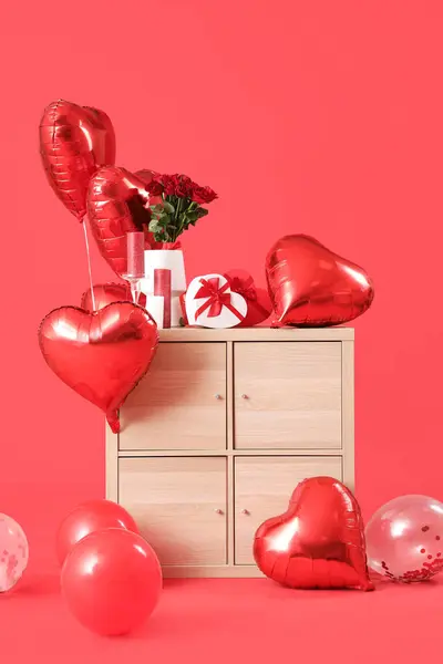 Wooden chest of drawers with heart-shaped air balloons and gifts on red background. Valentine\'s day celebration