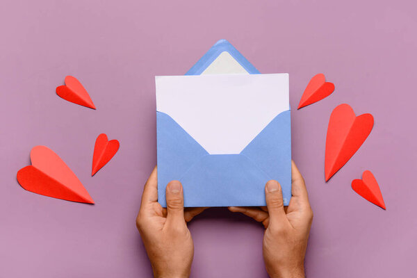 Male hands with envelope, blank card and red paper hearts on lilac background. Valentine's Day celebration