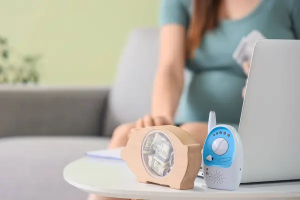 Piggy bank with baby monitor and laptop on table of pregnant woman at home, closeup. Maternal Benefit concept