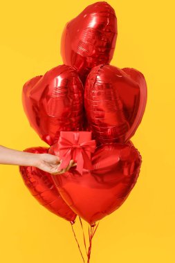 Hediye kutusu ve sarı arka planda kalp şeklinde balonları olan bir kadın eli. Sevgililer Günü kutlaması